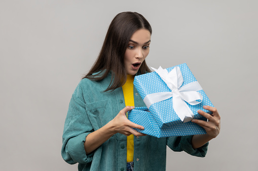 Astonished woman with dark hair unboxing blue gift box, looking inside with amazed facial expression, birthday present, wearing casual style jacket. Indoor studio shot isolated on gray background.