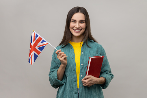 Portrait of attractive smart clever cheerful woman holding in hands book and british flag, education courses abroad, wearing casual style jacket. Indoor studio shot isolated on gray background.