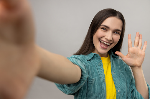 Positive optimistic woman taking selfie POV, flirting, expressing happiness, waving hand, saying hello, wearing casual style jacket. Indoor studio shot isolated on gray background.