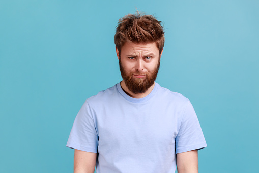 Portrait of bearded upset young man standing and looking at camera with dissatisfied sadness face, expressing sorrow, having bad mood. Indoor studio shot isolated on blue background.