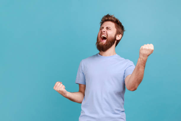Man with excited expression, raising fists, screaming, shouting yeah celebrating his victory success Portrait of overjoyed bearded man standing with excited expression, raising fists, screaming, shouting yeah, celebrating his victory, success. Indoor studio shot isolated on blue background. male likeness stock pictures, royalty-free photos & images
