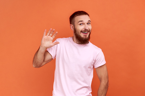 Portrait of positive handsome bearded man waving hand and friendly smiling at camera, making hi welcome gesture, wearing pink T-shirt. Indoor studio shot isolated on orange background.