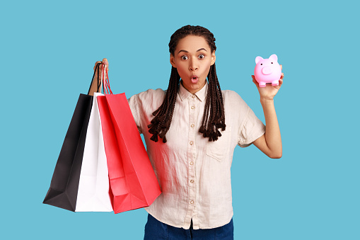 Astonished amazed woman with black dreadlocks holding shopping bags and piggy bank, cashback from buying purchases, wearing white shirt. Indoor studio shot isolated on blue background.
