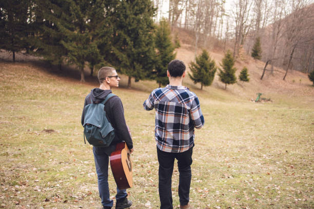 Two male friends going outdoors for fun in late autumn time, In this photo, they are walking in the park or forest with one of the holding a guitar on the move.