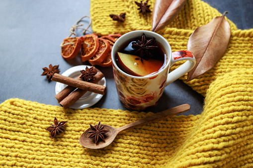 Istanbul, Turkey-March 4, 2022: Black tea with apple slices and star anise in a floral porcelain mug. There is a hand-knitted yellow scarf wrapped around the mug. And there are star anises, cinnamon sticks, yellowed leaves, dried tangerine slices. The table is dark gray. Shot with Canon EOS R5.