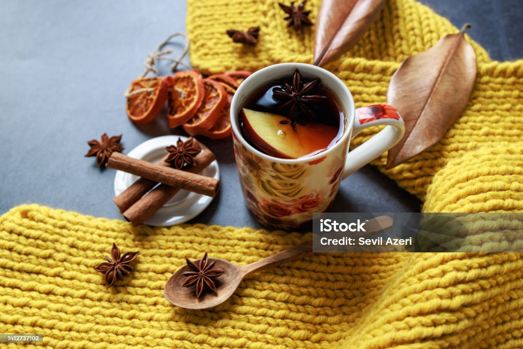 Black tea with apple slices and star anise in a floral porcelain mug Istanbul, Turkey-March 4, 2022: Black tea with apple slices and star anise in a floral porcelain mug. There is a hand-knitted yellow scarf wrapped around the mug. And there are star anises, cinnamon sticks, yellowed leaves, dried tangerine slices. The table is dark gray. Shot with Canon EOS R5. Tea - Hot Drink Stock Photo
