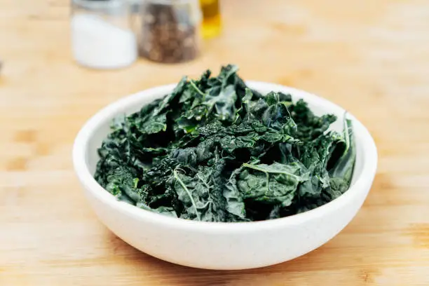 Photo of Teared curly green kale leaves in the plate with seasonings and olive oil on the kitchen table. Ingredients for kale chips or healthy salad meal. Healthy eating, dieting lifestyle. Selective focus