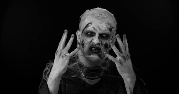 istock Scary wounded zombie undead guy making faces, looks at camera hiding through hands smiles terribly 1412746692