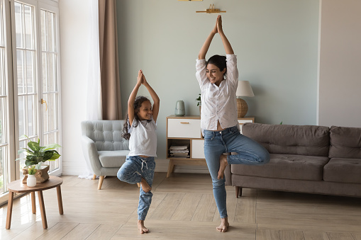 Happy Indian yogi mom teaching daughter kid to do yoga at home, standing in tree pose, smiling, laughing, talking, enjoying home exercises, leisure activities, keeping healthy life style