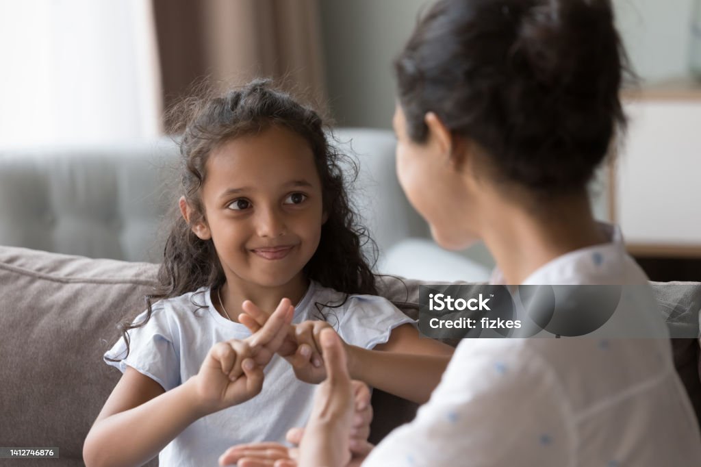 Happy cute Indian kid girl talking to mother, using hands Happy cute Indian kid girl talking to mother, using hands, fingers, speaking sign languages, sitting on sofa at home. Female teacher, therapist teaching child with deafness to communicate Child Stock Photo