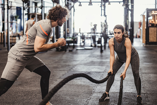 Young caucasian woman exercising by doing a battle rope in cross gym or fitness club. She crossing the rope with both hands. The man giving encourage beside the woman