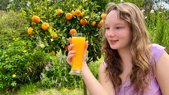 the girl drinks orange juice against the background of a tangerine tree, it can be orange juice tangerine mango she drinks greedily and really likes the juice delicious everywhere greens and summer.