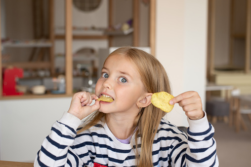 Caucasian little girl puts a snack in her mouth with her hand. The child sits at home at the table and enjoys eating fast food, potato chips. The concept of delicious and unhealthy food for children