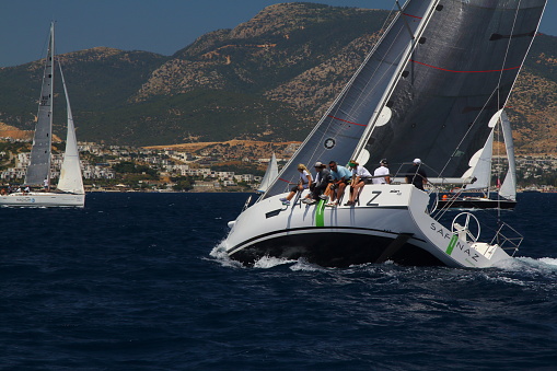 Bodrum,Mugla, Turkey. 26 May 2019: sailor team driving sail boat in motion, sailboat wheeling with water splashes, mountains and seascape on background. Sailboats sail in windy weather in the blue waters of the Aegean Sea, on the shores of the famous holiday destination Bodrum. Men and women on a sailboat in a very cold weather continue to cruise. Sailing crew on sailboat during regatta. Sailing in the wind through the waves at the Sea. Close up of sailing boat, sail boat or yacht at sea with white sails.