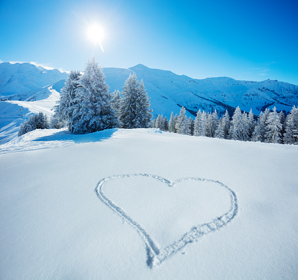Heart symbol drawn in the snow on top of the mountain slope with snowy firs and range tops on background