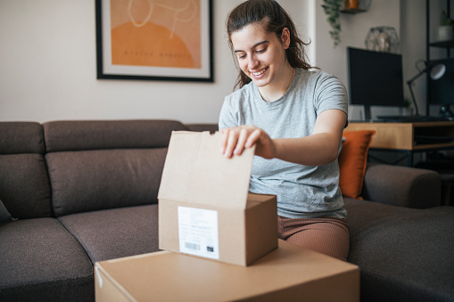 Smiling young woman sitting on the sofa and opening a box with delivered products.