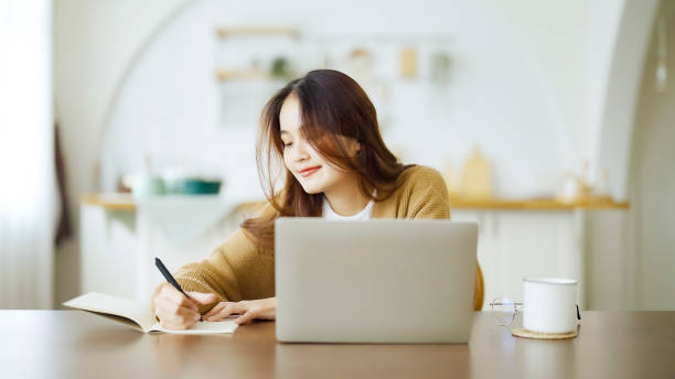 istock Young asian woman sitting at table and taking notes in notebook. Working on table with computer laptop, document paperwork and cup of coffee. Student learning online 1412782895