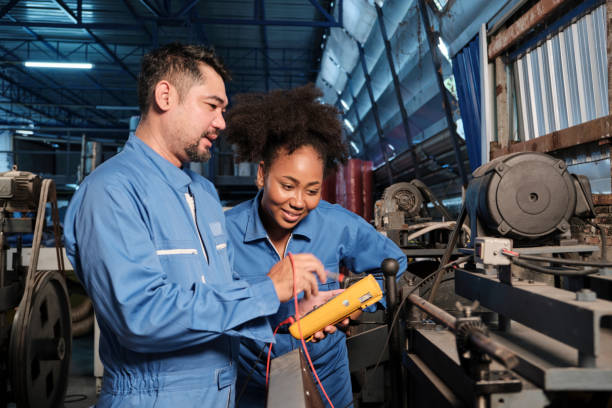 Two professional engineers inspect machines' electric systems at the factory. Asian male and female African American engineers in safety uniform work by inspecting machines' voltage current, checking, and maintaining at manufacture factory, electric system service occupations. dynamometer stock pictures, royalty-free photos & images