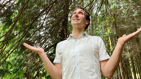 a handsome European man looks at the sky and raises his hands up he is in a white shirt and forest behind him. High quality 4k footage