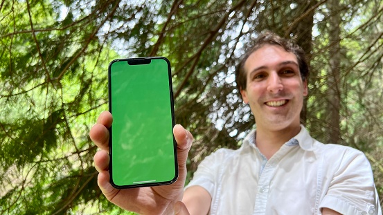 a handsome spanish man shows a phone on which a green screen chromakey he stands in a forest wearing a white shirt. High quality 4k footage