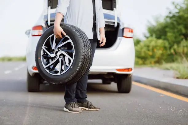 Handsome Asian young man holding spare wheel against for changing flat tire on the road.