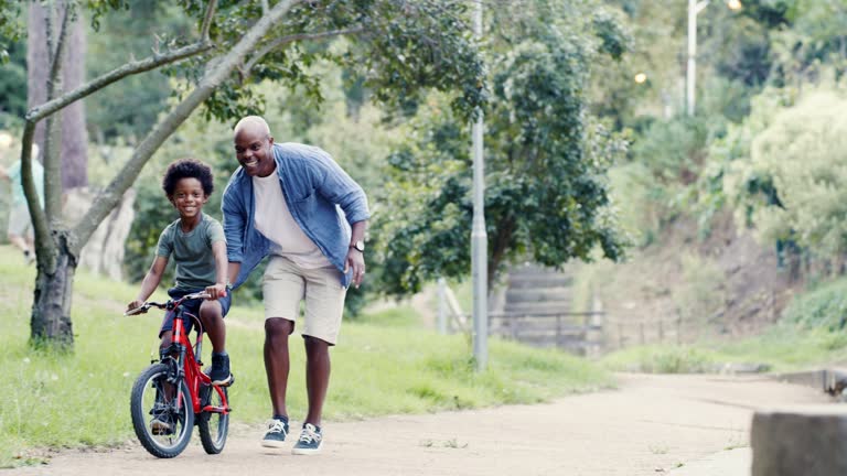 Happy, excited and playful boy being taught to ride a bike by his father in a nature park. A proud african american dad helping and teaching his little son to ride a bicycle outside in a forest