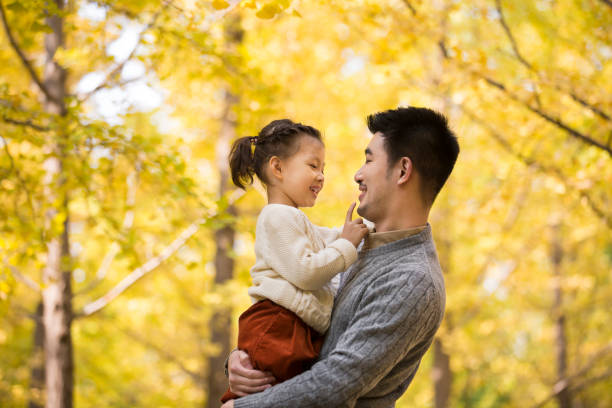 Young Chinese father holding his daughter in his arms in the autumn forest, the little girl's hand touching her father's scruff - stock photo stock photo