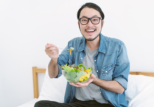 Healthy lifestyle young Asian bearded man eating fresh green salad from bowl smiling and looking at camera while sitting at home. Korean man like to eat vegetables, healthy food, vegetarian products.
