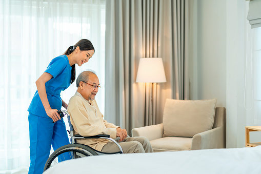 Female home caregiver pushing with senior man, sitting on the wheelchair in living room . Home healthcare concepts.