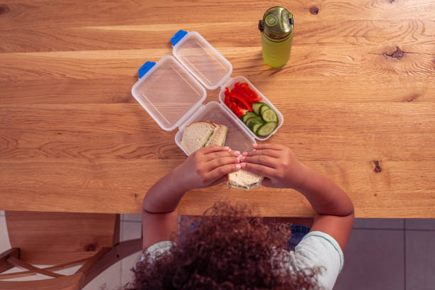 Top view image of a little student girl eating healthy lunch at school. Directly above image of a school girl eating healthy lunch during lunch break in classroom. bottled water on table stock pictures, royalty-free photos & images