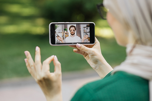 Young muslim woman, resting at park with digital smart phone, having a video call with her friend, handsome bearded man, showing his care routine with eye patches.