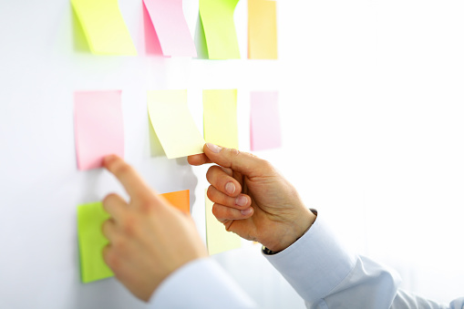 Close-up of man picking up yellow and pink lists with empty space. Persons hands and white board. Businessman in suit. Light personal cabinet. Office and business concept