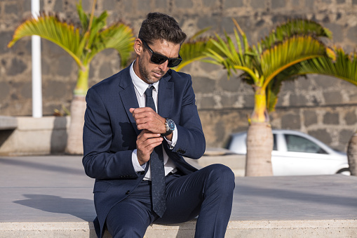 Serious male entrepreneur in suit and sunglasses fastening expensive wristwatch while sitting on step against palms and stone wall on city street