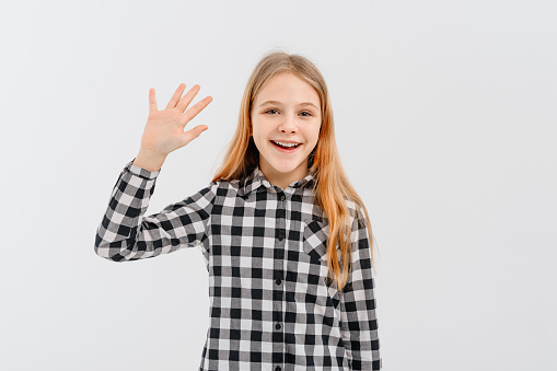 A friendly teen girl says hello, waves hand and smiles happy at you, stands over white background in casual shirt
