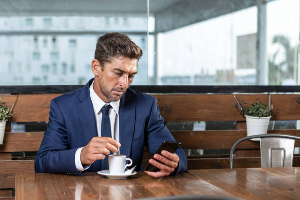 Businessman using smartphone in cafe Male entrepreneur in suit stirring coffee in cup and browsing social media on cellphone while sitting at table on cafeteria terrace stirring stock pictures, royalty-free photos & images