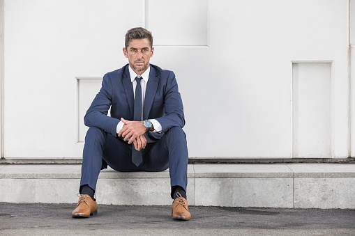Full body male executive in suit sitting on marble border and looking at camera against white wall while resting on city street after work