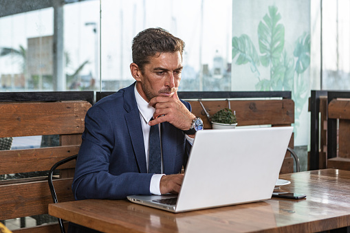Pensive male entrepreneur in suit touching chin and thinking over data on netbook while sitting at table and working on business project in cafe