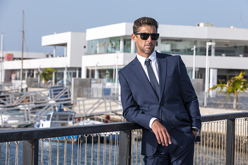 Serious businessman in suit and sunglasses leaning on bridge banister and looking away against modern building on sunny day in city harbor