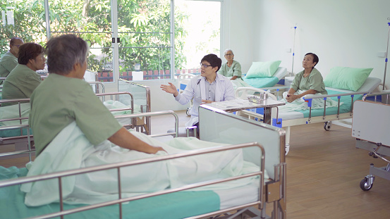 An Asian doctor talking to group of old elderly patient or pensioner talking, have a meeting, in nursing home in hospital. Senior people lifestyle activity recreation. Health care physical therapy.
