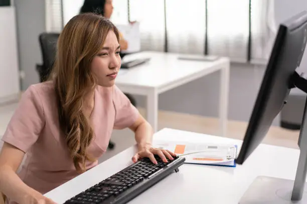 Photo of Asian female employee having farsighted problem looking at monitor, Blue light hazard from computer screen. woman tired of focusing on reading and working in the office.