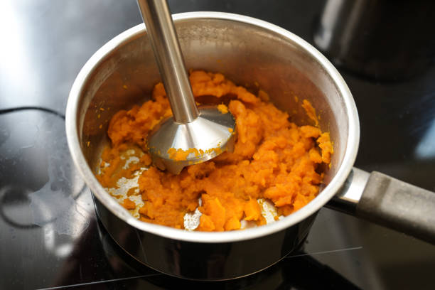 le carote cotte vengono frullate con un frullatore in una pentola d'acciaio su una stufa nera per un pasto vegetale sano e digeribile o una zuppa contro la diarrea, focus selezionato - blender concepts red black foto e immagini stock