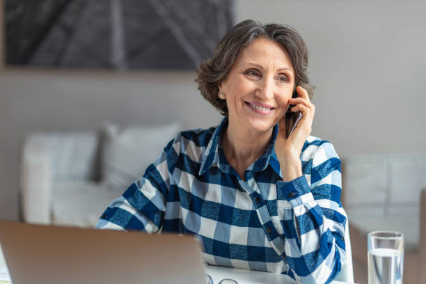 istock Beautiful senior woman working talking on mobile phone sitting at the workplace with computer laptop in a modern office or home, online remote work, smiling 1412837243