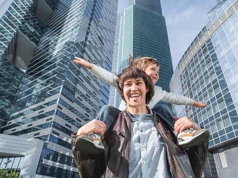 Laughing boy plays like airplane on father's shoulders. Dad and son looks on glass walls of buildings. Future and modern technologies, life balance and family life in well keeps districts.
