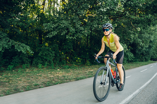 Young woman cyclist riding road bicycle on a free road in the forest at a sunny day. Healthy lifestyle concept. Wearing active wear and a protective helmet. Copy space.