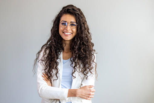 Waist up portrait modern business woman in the office with copy space. Portrait of a happy and confident young woman standing posing against a gray wall. Businesswoman with arms crossed looking at camera