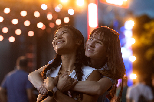 Two Asian and Caucasian girls hug and enjoy the atmosphere under the stage spotlights at a music festival.