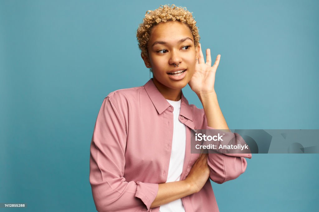 Curious African American lady in yellow summer clothes holding hand over ear trying to hear more, listening gossips, rumors, looking at camera with concentrated face expression. Body language concept Cute curious African American girl in pink casual clothes holding hand over ear trying to hear more, listening to rumors, gossips standing against blue wall with concentrated facial expression Gesturing Stock Photo