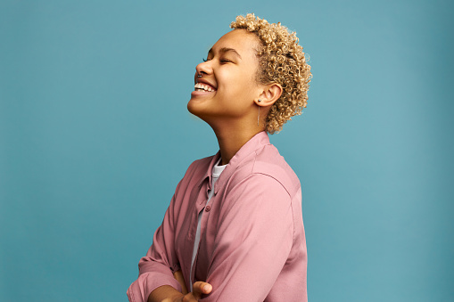 Profile view of happy joyful carefree female with dark skin and blonde curly hair, standing against blue background in pink shirt with arms folded, putting head up. Human emotions and feelings concept