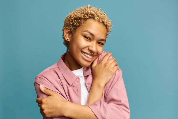 istock Charming gorgeous Afro woman keeps eyes closed, smiles with pleasure, shows white teeth feels comfort, hugs herself, wears orange jumper, tilts head, models over blue background, has high self esteem 1412855413