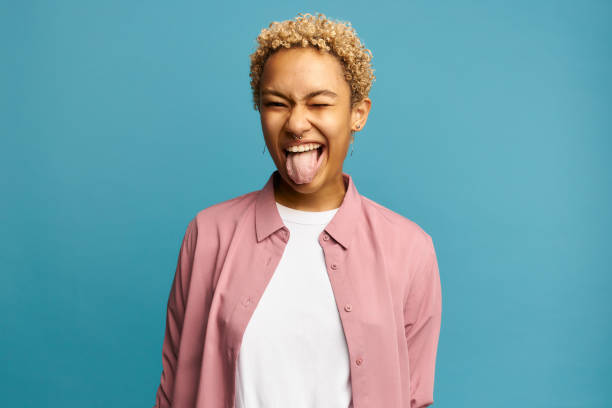 istock Pretty joyful African American with cute short curls, making faces in front of camera showing her tongue, after hitting wrist, having cheerful and carefree look, posing against orange wall in crop top 1412855476